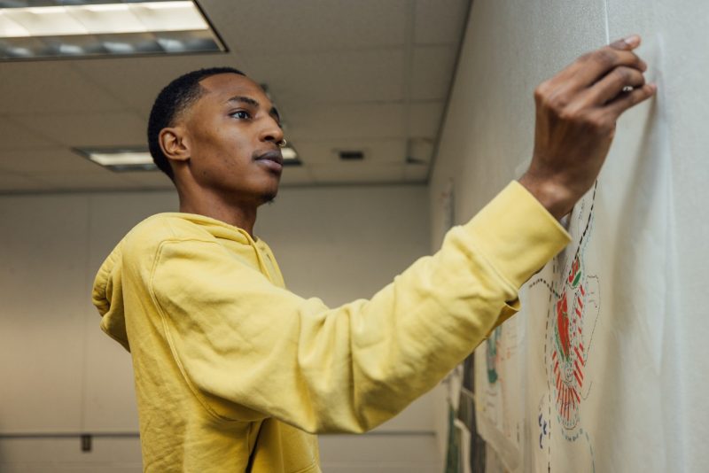 A Black male student in a yellow hoodie pins up landscape architecture designs on a wall in Cowgill Hall.