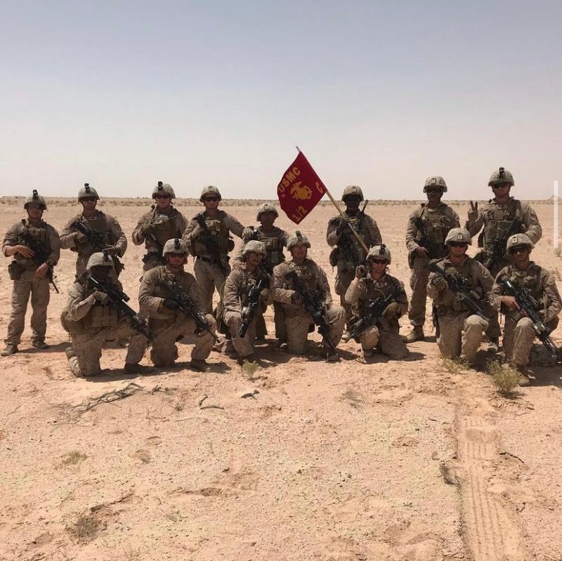 Fifteen Marines in camoflauge, holding guns, gather around a Marine Corps flag in a desert landscape..