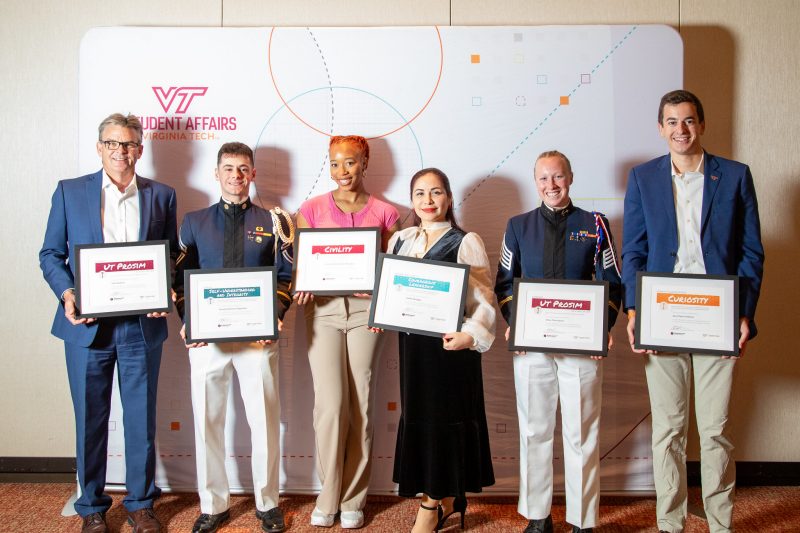 Five students and one faculty member pose with their framed awards in front of a Student Affairs background.