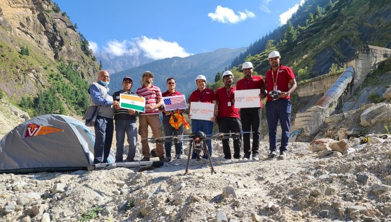 A group of 8 men stand in front of tall mountains and behind a large drone, holding up Indian and American flags and Virginia Tech signs.