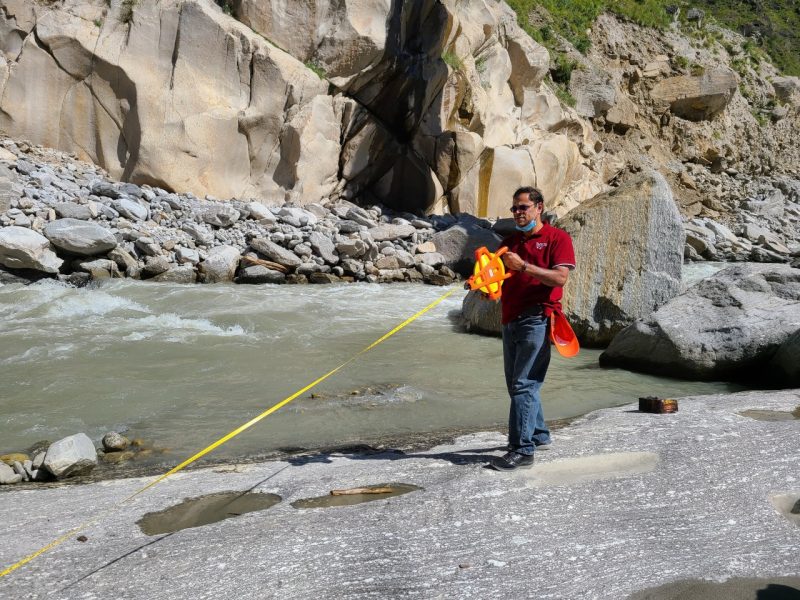 A man stands on rocks in front of a river, using a measuring device.