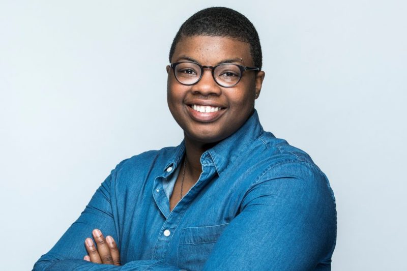 A young woman wearing glasses and a blue button-up shirt crosses her arms and smiles for a photo.