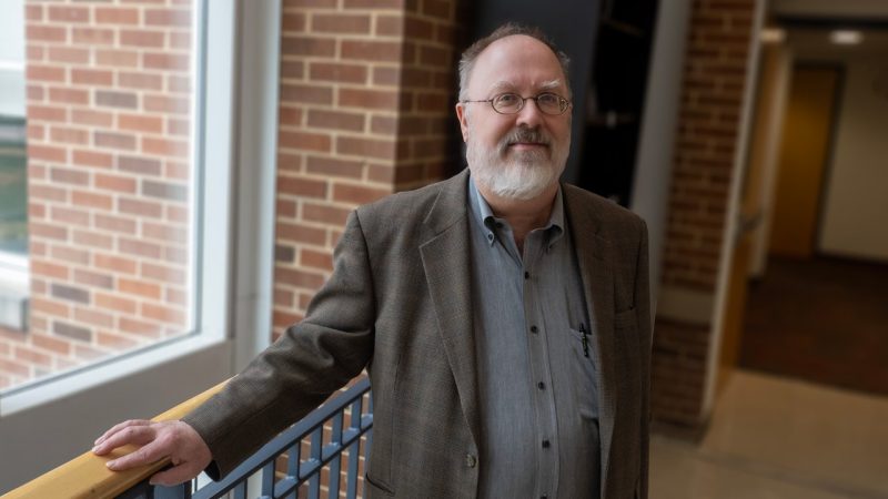 Joseph Eska 2024 Mid-length portrait of a man with a gray beard, wearing glasses, standing between two brick columns.