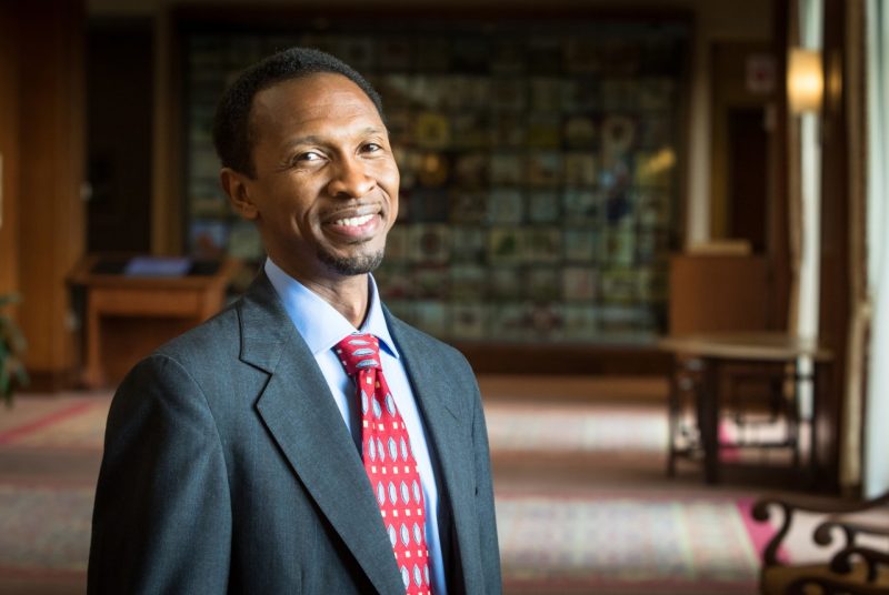 Sylvester Johnson, dressed in a gray suit with a blue shirt and red tie, stands to the left of the frame and smiles for a photo.