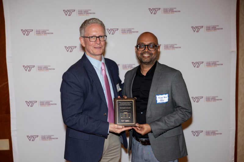 Two men stand and pose with the man on the right holding an award.