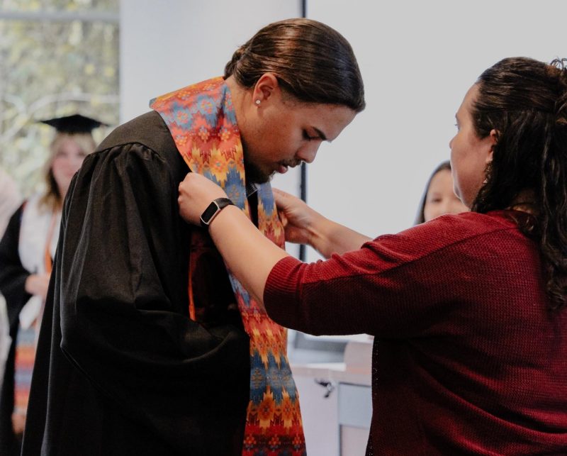 A student in graduation garb is bent forward, receiving a colorful stole from a community member. They are placing the stole around the back of the student's neck.
