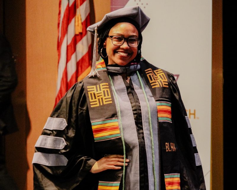 Posed student with stole and doctoral cap at the Donning of the Kente event Spring 2024 A young student poses with their hand on their hip and smiling into the camera, wearing the Kente stole and a doctoral cap.