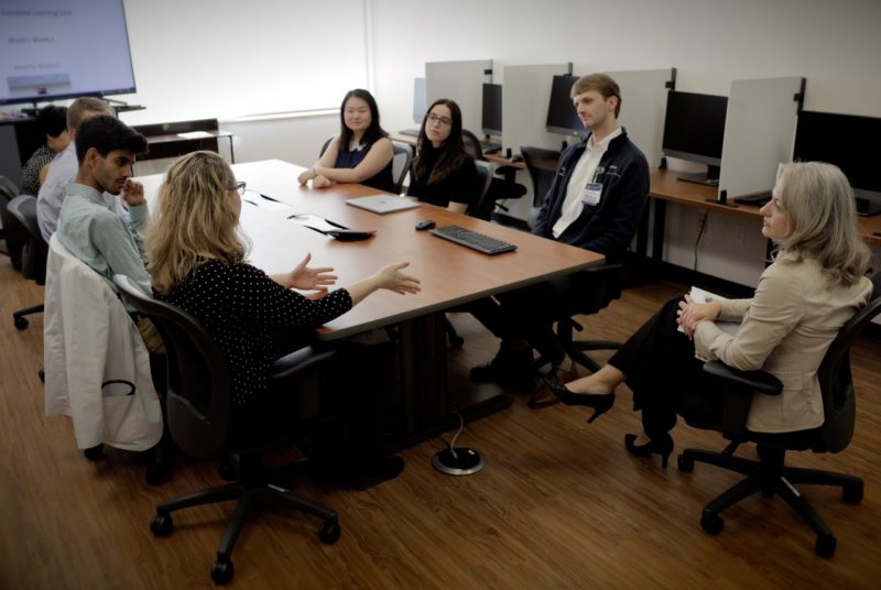 Seven students sitting around a table engaging in animated discussion. Woman observing.