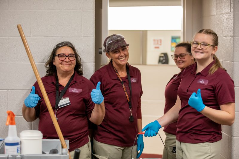 Four people smile at the camera while holding a "thumbs up" at the camera. The employees wear blue gloves and hold cleaning supplies.