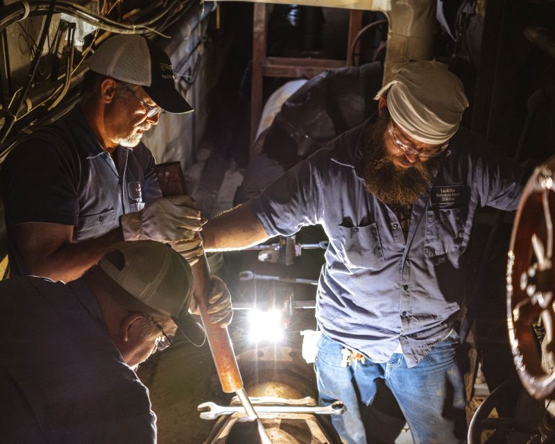 Three people wearing uniform shirts, hats, and jeans use tools to perform maintenance on a steam distribution pipe inside a steam tunnel.
