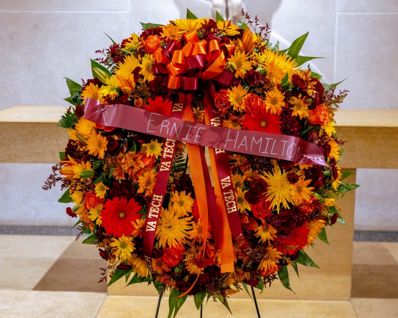A wreath filled with orange, yellow, and red flowers is placed in War Memorial Chapel. A banner across the wreath says, "Ernie Hamilton."