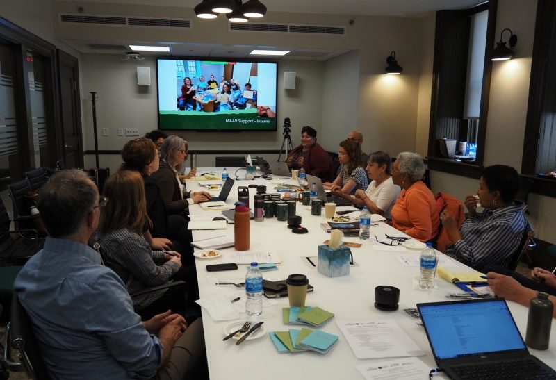 A group of team members with the project sit at a long table in a conference room while other partners involved with the project appear in a video conference call on a TV screen