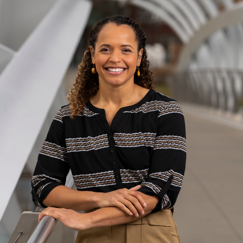 Sydney Turner A headshot of Sydney Turner standing with her elbow leaning on the railing of a large bridge, the background blurred behind her. She is wearing a striped top and has her tight curls pulled back in a half-up, half-down hairstyle.