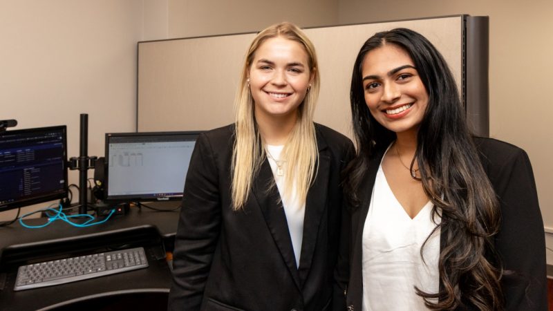 Virginia Intern Day Two young women dressed in black blazers and white shirts stand in front of a computer in an office.
