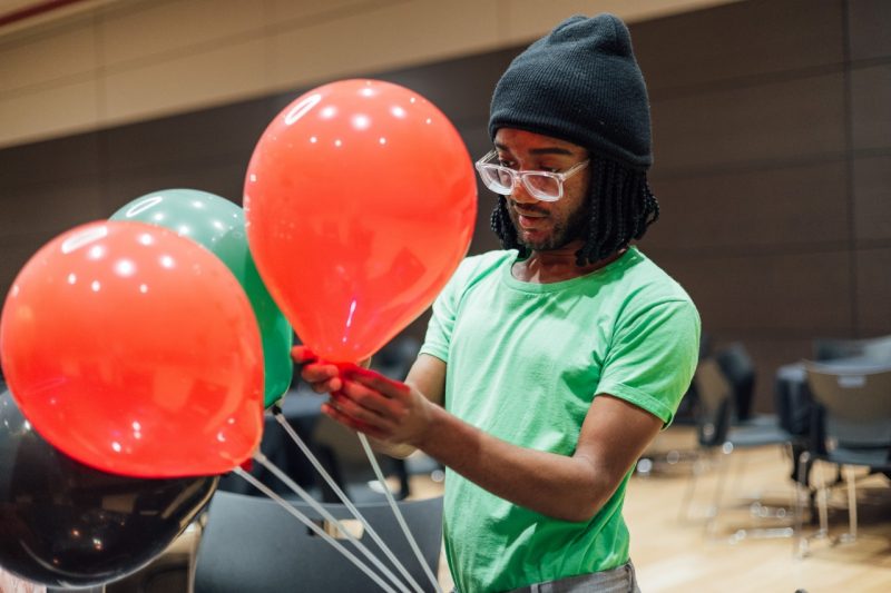 Renaldo Lynch, in a green t-shirt and black beanie, arranges orange and green balloons for a Ujima celebration in Squires.