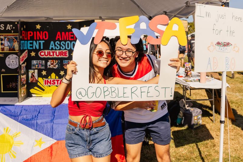 Two students in summer dress hold up a paper frame around their faces that says "VT FASA @GOBBLERFEST!" in multiple pastel colors. Behind them is a table with the national flag of the Philippines and multiple cardboard displays with photographs of students partaking in FASA's activities. A white pop-up tent is above them.