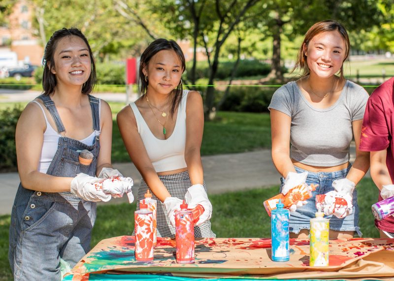 Three female students are outdoors, one dressed in overalls, another in a white tank-top and patterned skirt, and the last in a grey t-shirt and denim shorts, apply bottles of dye to white fabric outdoors. The table in front of them is covered with color-stained brown paper and holds other bottles of dye. Several green trees are out of focus behind them.