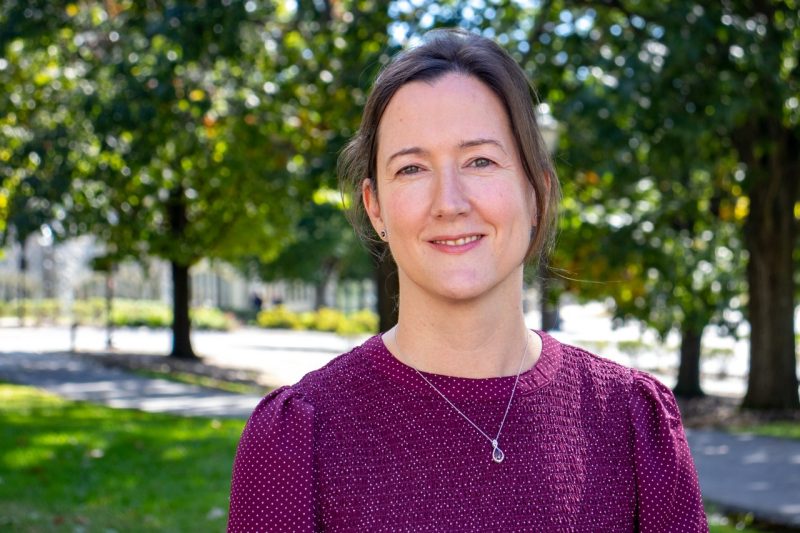 A smiling woman wearing a purple blouse stands in front of trees on a sunny day.