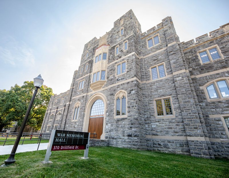 The sun shines behind Grey Hokie Stone War Memorial Hall on a clear day.