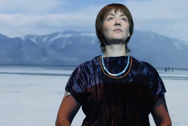 Amelia WInger-Bearskin - a white woman with short dark hair, wearing a dark purple and black top with beaded necklaces - looks up, light shining on her face, as she stands in front of an image of snowy mountains and gray skies.