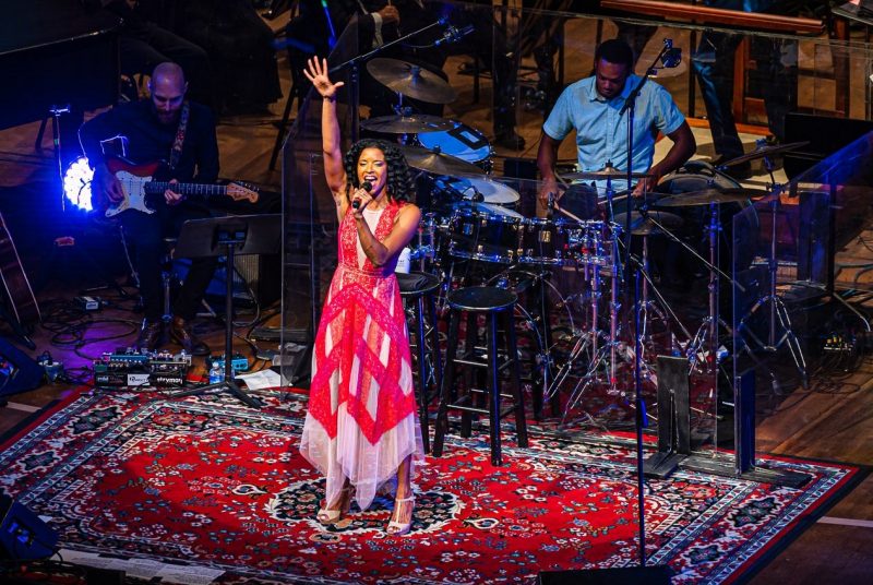 Broadway star Renée Elise Goldsberry, a young Black woman, belts out a note on stage in a long pink dress, her arm in the air above her head, as an orchestra performs behind her.