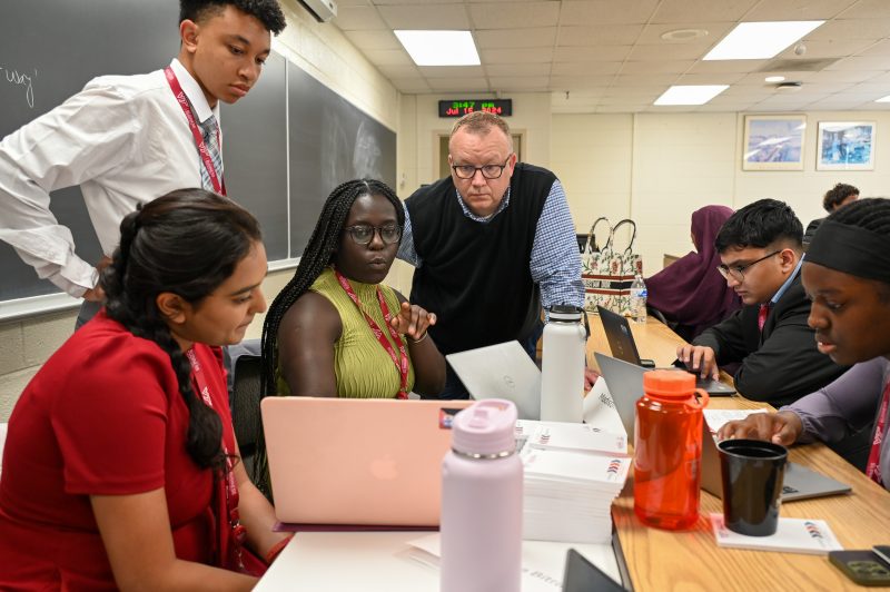 Group of students working on laptops at a table while Ron Poff looks on.