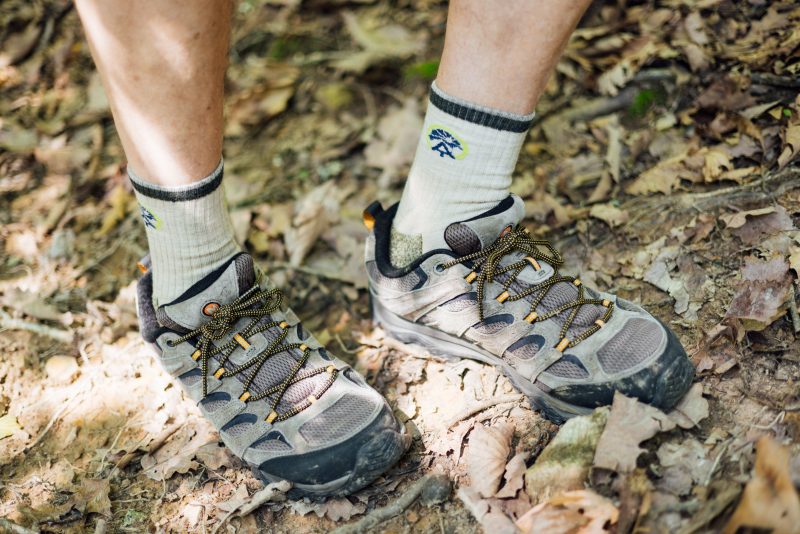 Don's hiking shoes and socks A close-up photo of Don Taylor's feet wearing well-worn hiking shoes and light-colored socks, standing on a forest floor covered with dry leaves and twigs. Don's crew socks feature the AT logo on the ankles.