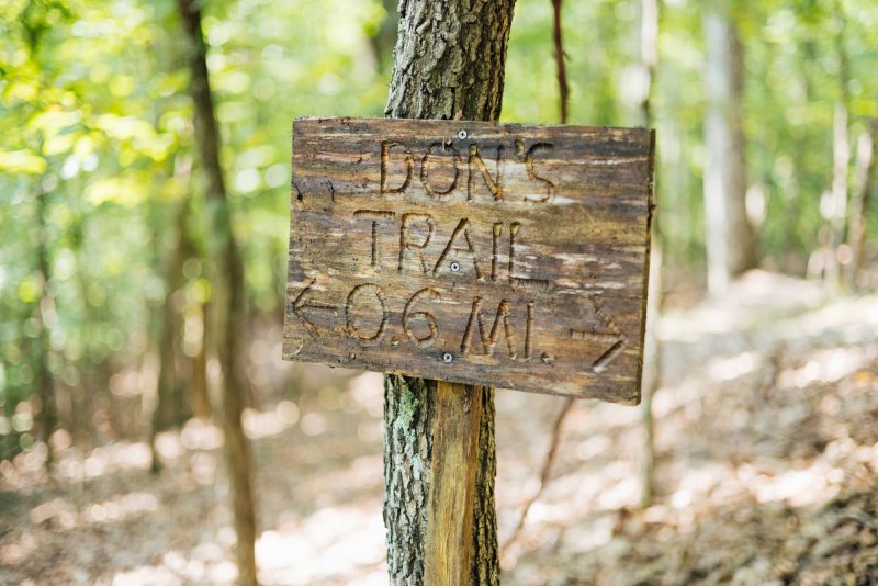 Don Taylor's trailhead sign The image shows a wooden sign that Don made for the trailhead of the trail he built on his property behind his house. It's weathered, leaning against a tree and is carved with the words "Don's Trail, 0.6 MI" with an arrow pointing left and an arrow pointing right to indicate the trail is a loop. The background is softly blurred, revealing a densely wooded area with green foliage and a serene, natural atmosphere. The sign and the surroundings suggest a well-worn, personal trail in the woods.