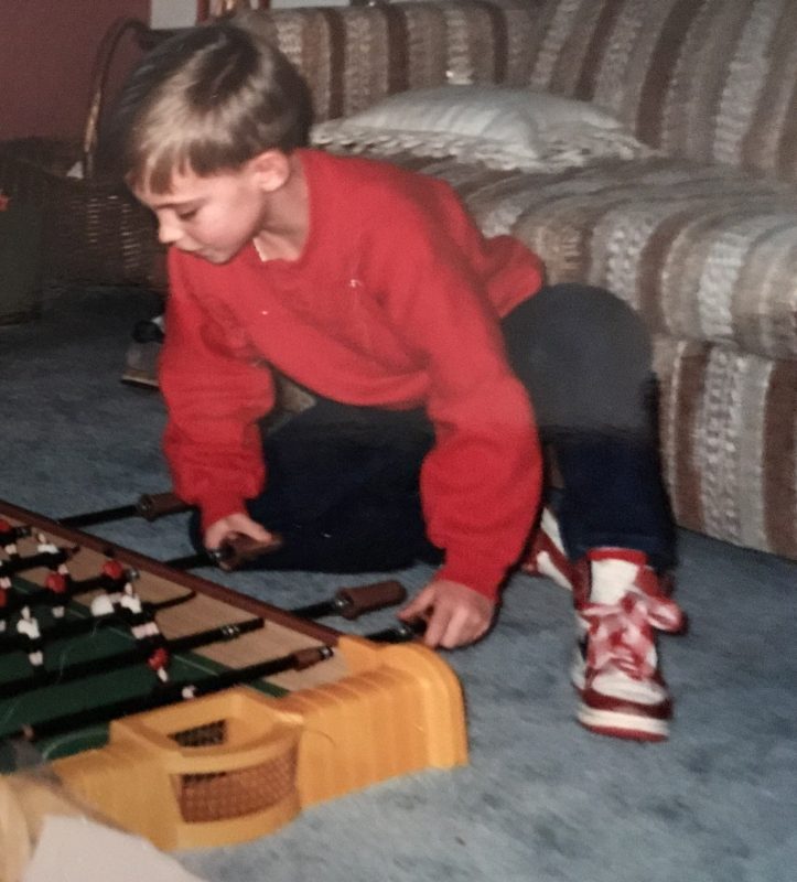 Jacob Barney childhood photo The vintage image shows a young Jacob Barney, kneeling on the floor while intensely focused on playing a tabletop foosball game. He is wearing a bright red sweater, dark pants, and red and white sneakers. The background includes a striped sofa and a wicker basket. The boy’s expression and posture convey concentration and engagement in his play, capturing a nostalgic and playful moment from childhood.