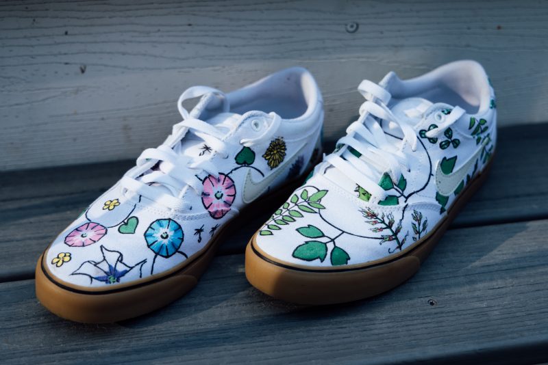 Jacob Barney's favorite sneakers The image is a close-up of a pair of Jacob Barney's white canvas Nike sneakers with custom hand-painted designs featuring various colorful botanical elements. The shoes are decorated with illustrations of flowers, leaves, and vines in shades of green, pink, blue, and yellow, creating a vibrant and artistic look. The sneakers are placed on a wooden surface, and the gum-colored soles contrast with the white canvas.