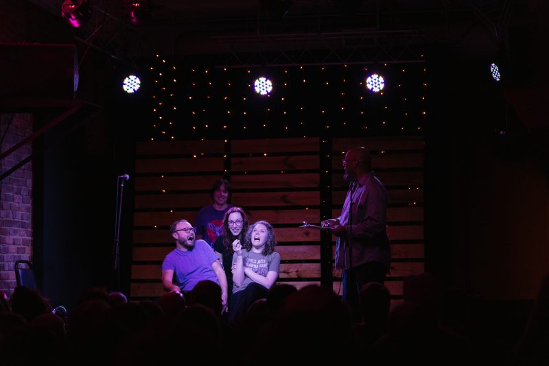 Sarah Parker on stage 2 The image captures a lively scene on stage at a small venue, where a group of four people is gathered under soft, colorful stage lighting performing an improv bit. In the foreground, Sarah Parker and two others are seated and sharing a humorous moment, laughing openly. The person on the left, wearing glasses and a blue shirt, is leaning back, laughing heartily. Sarah Parker is in the center, wearing a gray T-shirt with the text "GIRLS JUST WANNA HAVE FUNDING FOR SCIENTIFIC RESEARCH," is also laughing with her head tilted back. A third person, wearing a purple "Big Thief" T-shirt, stands behind them, smiling. On the right, a man is standing at a microphone, holding a clipboard and engaging with the group. The background features a backdrop of wooden panels and small string lights, adding to the warm and intimate atmosphere of the event. The audience's heads are visible in the foreground.