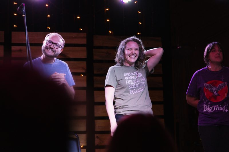 Sarah Parker on stage 3 The image shows Sarah Parker and two of her cast mates standing on a stage under soft lighting, with a backdrop of wooden panels and string lights. Sarah is in the center, smiling broadly while holding one hand behind her head. She is wearing a light green T-shirt with the text "GIRLS JUST WANNA HAVE FUNDING FOR SCIENTIFIC RESEARCH." To her left, a man with glasses is smiling warmly, clasping his hands together. He is wearing a striped T-shirt. On the right side, another woman, who is partially visible, is wearing a purple T-shirt with the "Big Thief" logo. The atmosphere is casual and joyful.