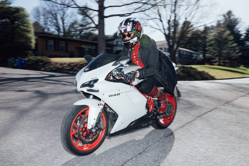The image shows Margie Lee riding a white Ducati 848 EVO motorcycle with bright red wheels. She is dressed in a distinctive outfit that includes a black coat, red boots, and gloves. She is also wearing a black helmet with a white and red design, along with sunglasses. The motorcycle is in motion as she rides down a residential street with houses and trees in the background. The overall setting suggests a brisk ride on a cool, clear day.