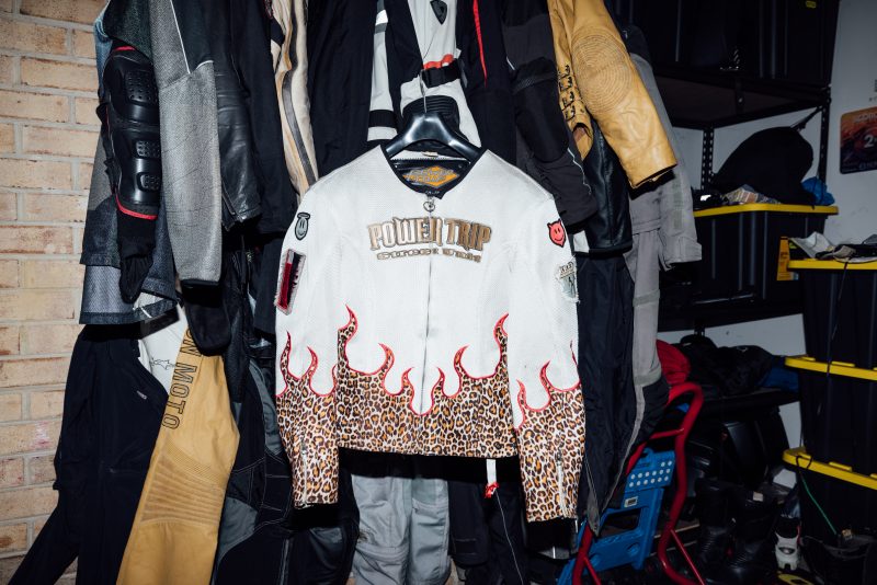 Margie Lee's motorcycle racing jacket The image shows a white motorcycle racing jacket with distinctive leopard print and red flame designs hanging on a hanger in a garage filled with various motorcycle gear. The jacket features the words "POWER TRIP" and "STREET UNIT" prominently displayed on the front, along with several patches on the sleeves. Surrounding the jacket are other motorcycle jackets and protective gear in various colors, including black, gray, and tan.