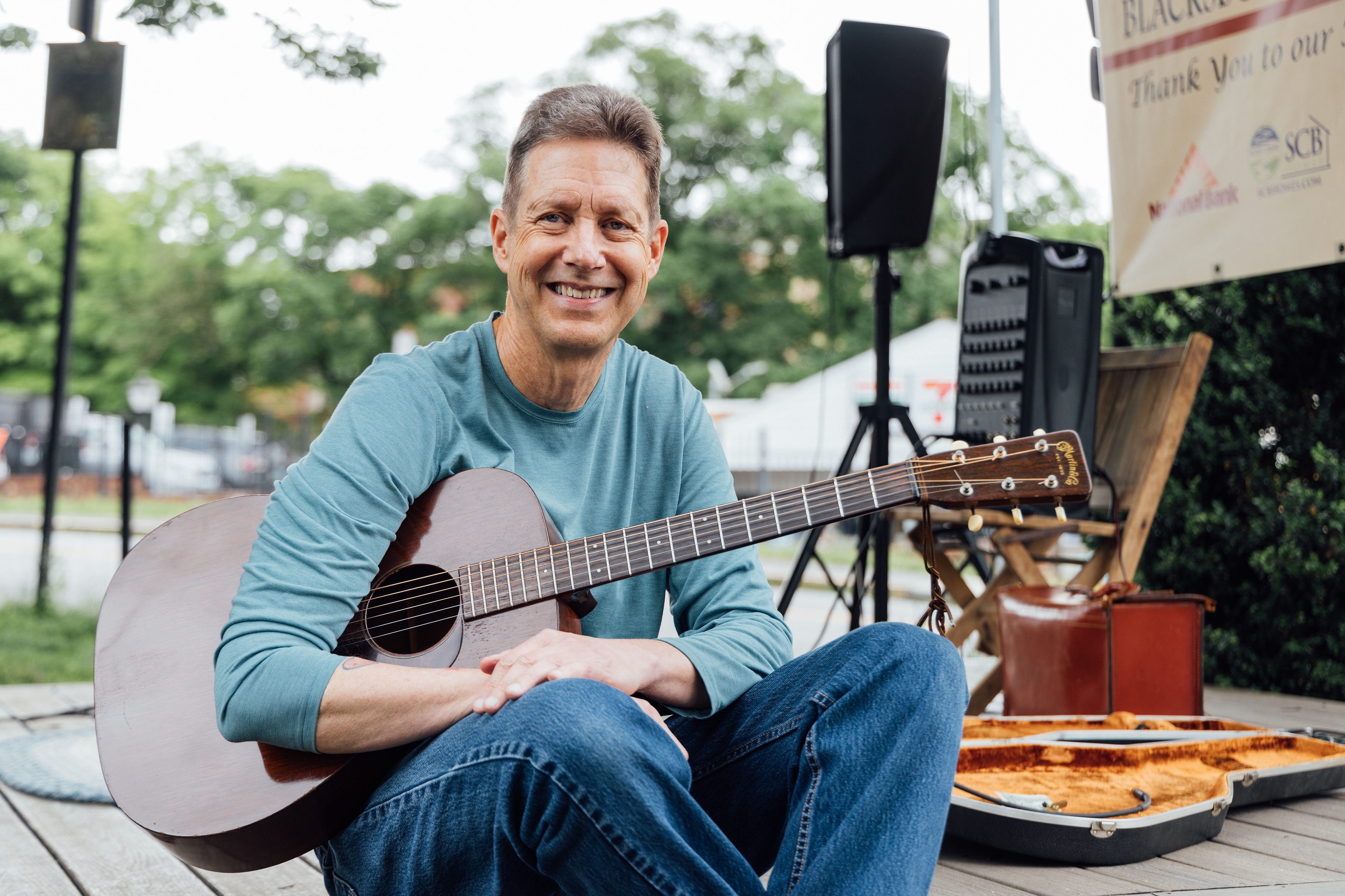 Paul Heilker The image is a portrait of Paul Heilker sitting outdoors on a wooden deck at the farmers market, holding an acoustic guitar. He is smiling warmly at the camera, creating a friendly and relaxed atmosphere. The man is wearing a teal long-sleeve shirt and blue jeans. The background shows some outdoor equipment, including a speaker on a stand, a guitar case, and some signage. The setting is in a green, tree-filled area with a casual and inviting vibe.