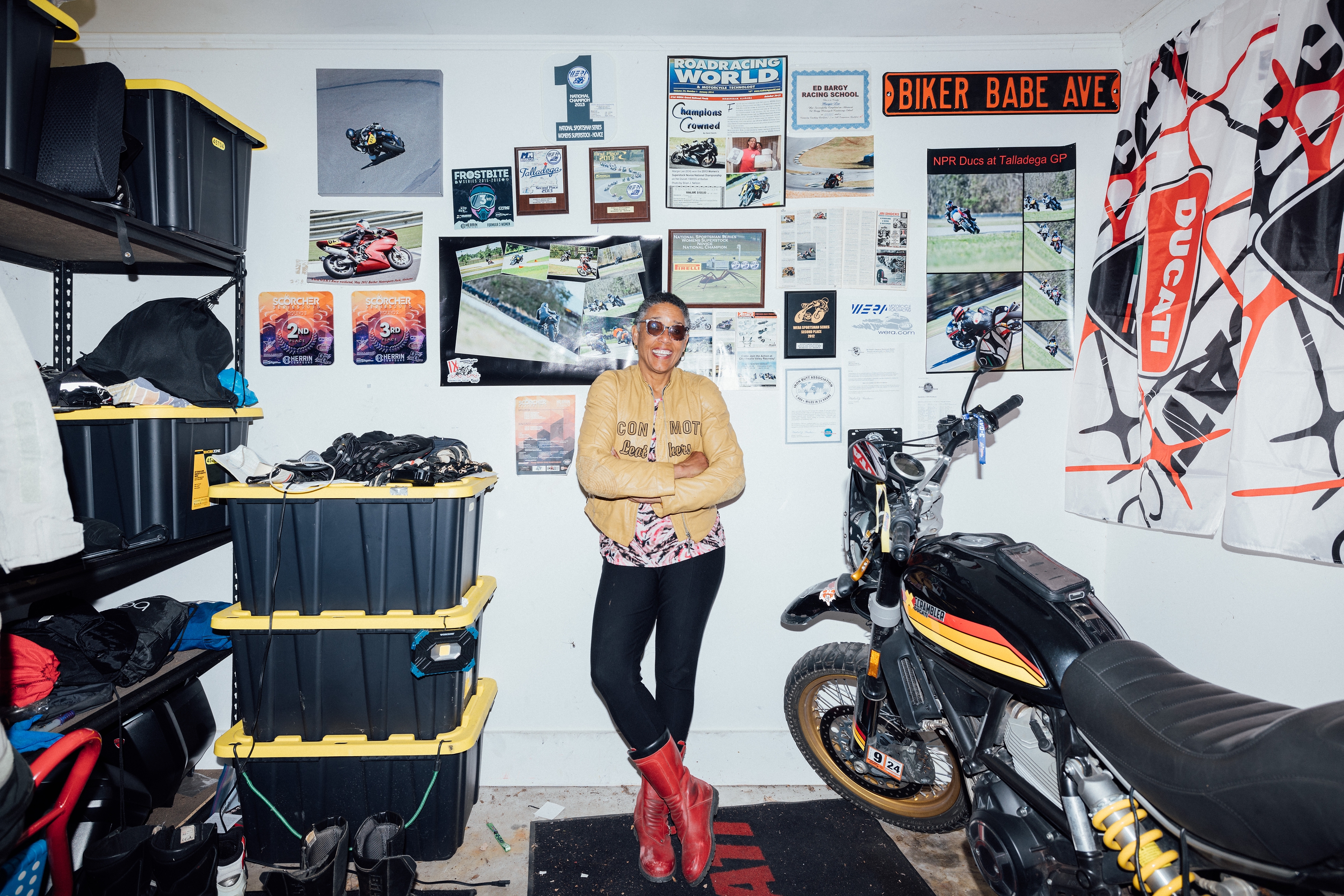 Margie Lee in her garage The image shows Margie Lee wearing a tan leather jacket, black pants, and red boots stands confidently in a garage filled with motorcycle gear and memorabilia. She is smiling with her arms crossed in front of a wall covered with racing photos, posters, and awards. To her right is a black and yellow racing motorcycle, and the room is organized with shelves holding plastic storage bins and various riding accessories. A sign on the wall reads 'BIKER BABE AVE,' adding to the theme of her passion for motorcycles.