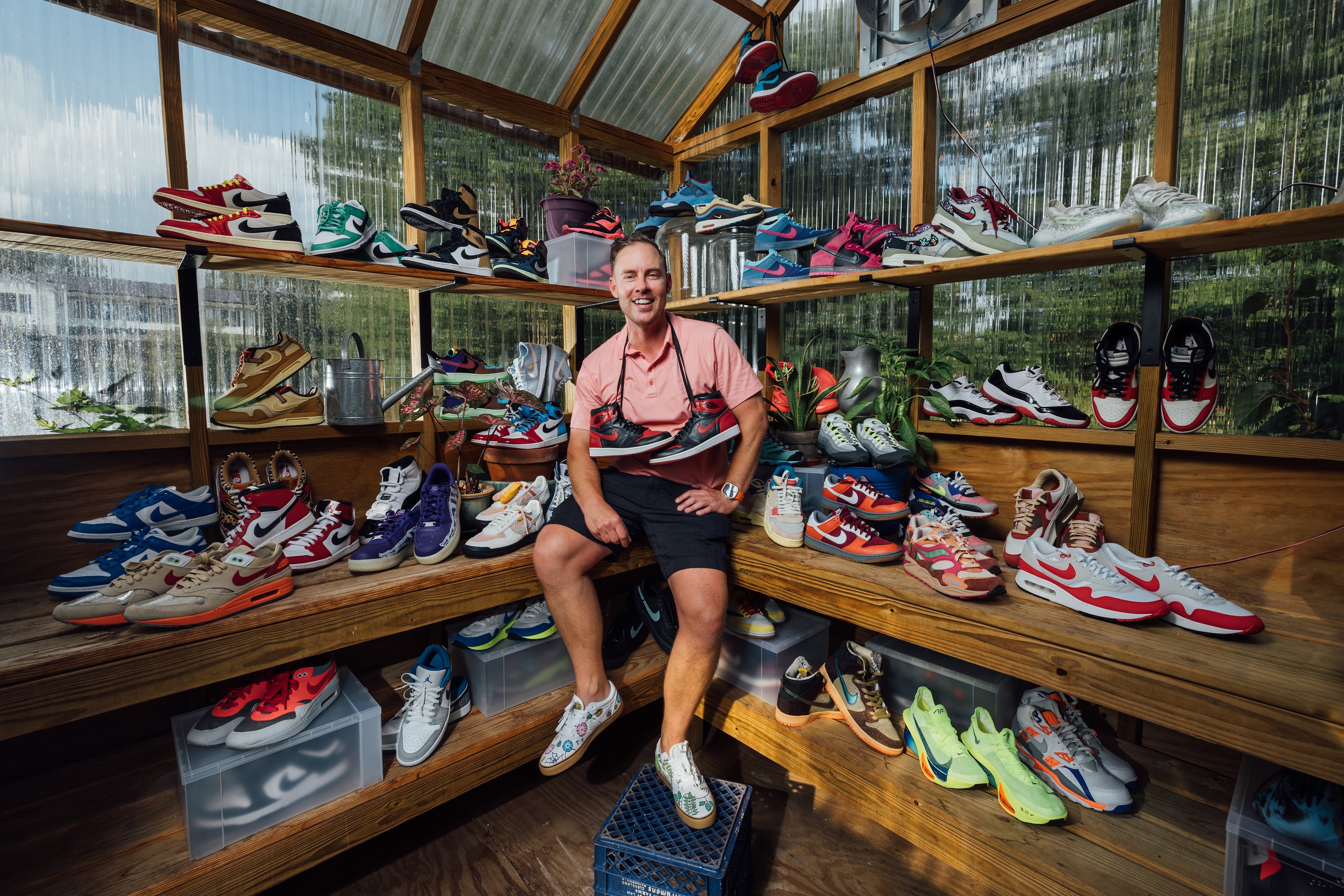 The image shows Jacob Barney wearing a light pink shirt and dark shorts sits in a wooden greenhouse surrounded by an extensive collection of colorful Nike sneakers. He is smiling and has a pair of red and black Air Jordans draped from his neck. The shelves around him are filled with a variety of sneakers in different styles and colors, neatly arranged, with some potted plants adding greenery to the scene.