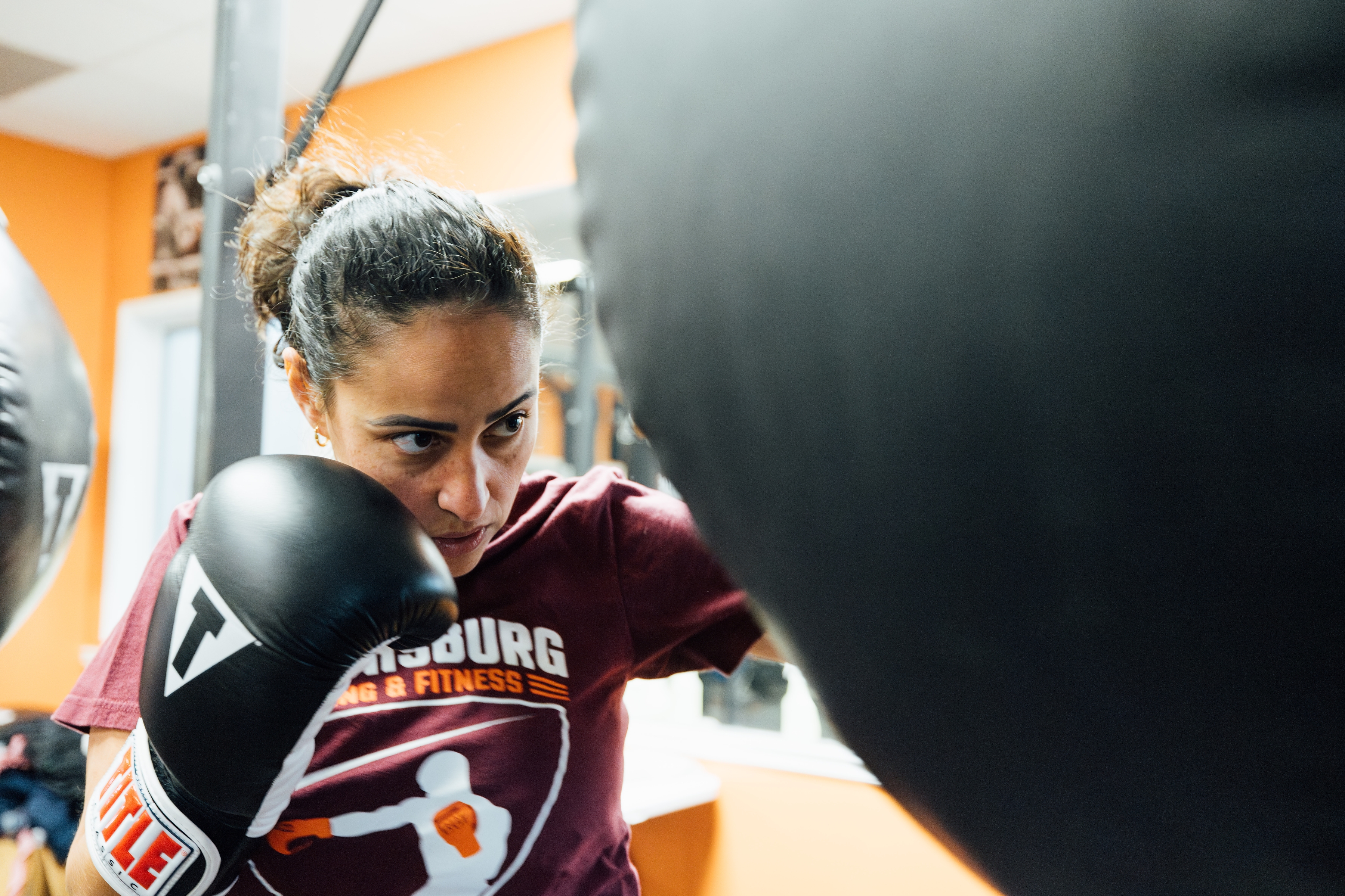 Renata Carneiro The image shows Renata Carneiro intensely focused during a boxing workout. She is wearing boxing gloves and a maroon T-shirt with the logo "Blacksburg Boxing & Fitness" on the front. She is throwing a punch towards a black punching bag, her expression determined and concentrated. The background shows a gym setting with orange and white walls.