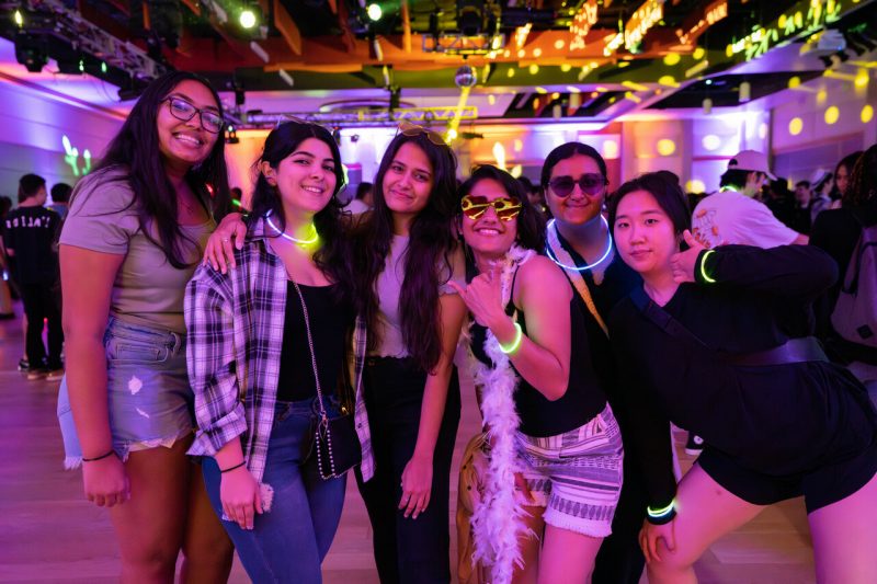 Six students smile in a disco dance hall, wearing glowing accessories.