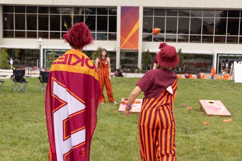 two students are standing with their backs to the camera, one wearing a Hokie flag as a cape and the other in orange and maroon striped overalls. Two students stand in front of them, and a beanbag is captured in the air mid-toss. The facade of Dietrick Hall is in the background behind them.