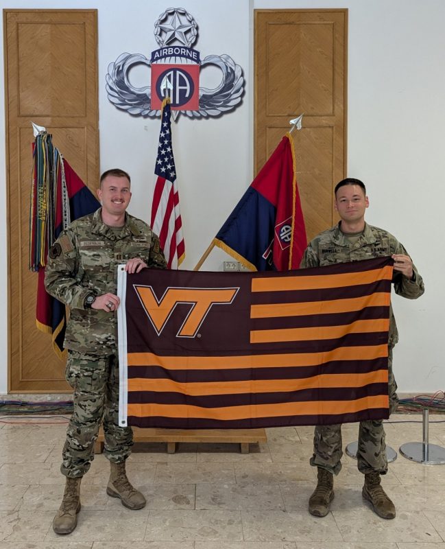  Two service members smile at the camera while holding a striped Virginia Tech flag. They are in camouflage uniforms. 