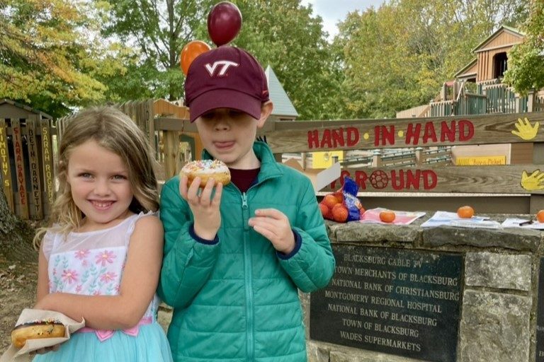 Two children with donuts stand in front of Hand-in-hand playground in Blacksburg. One child wears a VT cap.