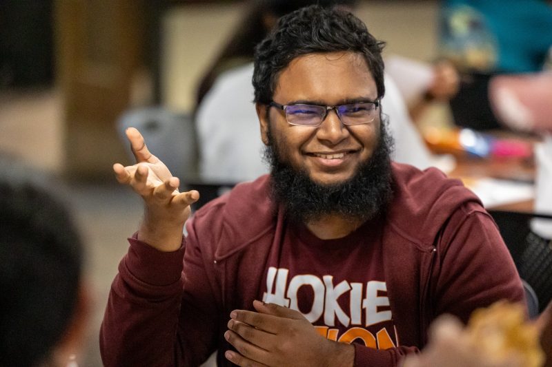 A student with a dark beard and wearing a maroon shirt with the word "Hokies" visible in an orange, bold font smiles and gestures as he engages in conversation at an Interfaith Initiative event.