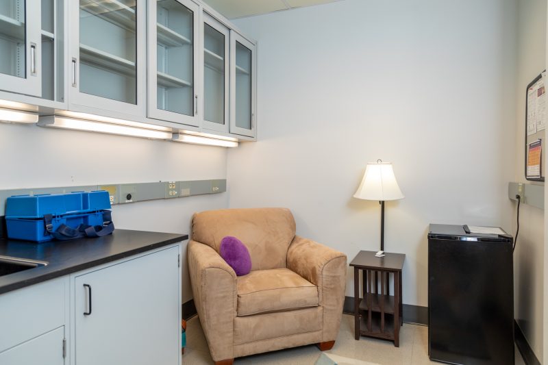 A lactation room with a tan armchair positioned next to an electrical outlet. The room also contains a mini fridge, a lamp, and a countertop.