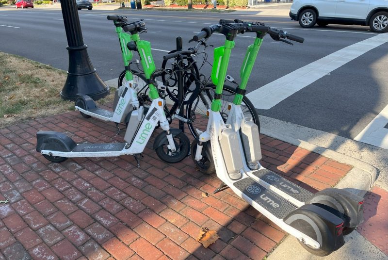 Tangled Scooters on Sidewalk Four electronic scooters are secured to a pole at the end of a sidewalk along a city street.