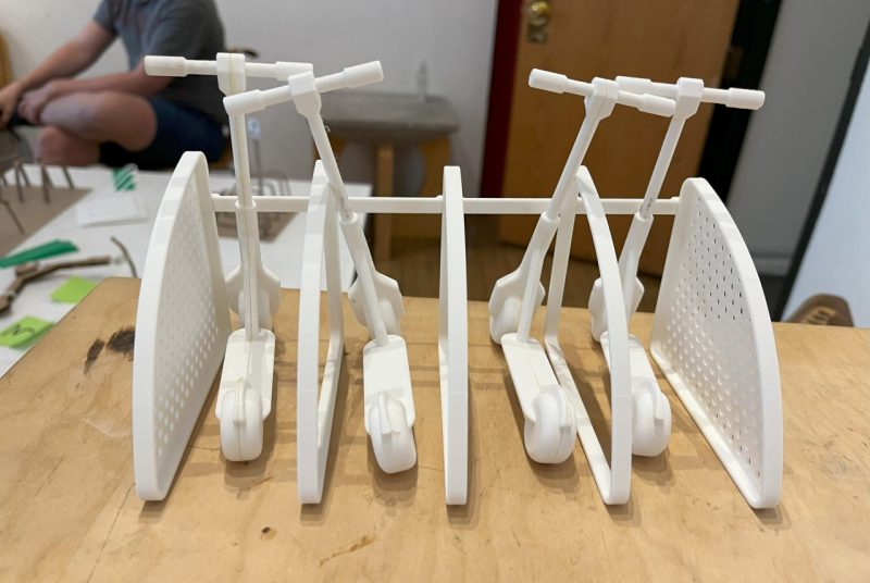 Scooter Rack Prototype A small plastic model of a scooter rack consisting of triangular sides and middle connected by horizontal bars, with four scooters resting on the bars near the ground.
