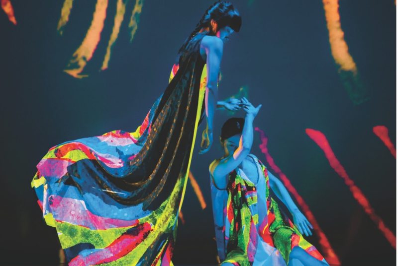 Two dancers with Cloud Gate Dance Theatre of Taiwan perform on stage wearing neon costumes that glows under a black light. One dancer sits while the other stands above her looking down.