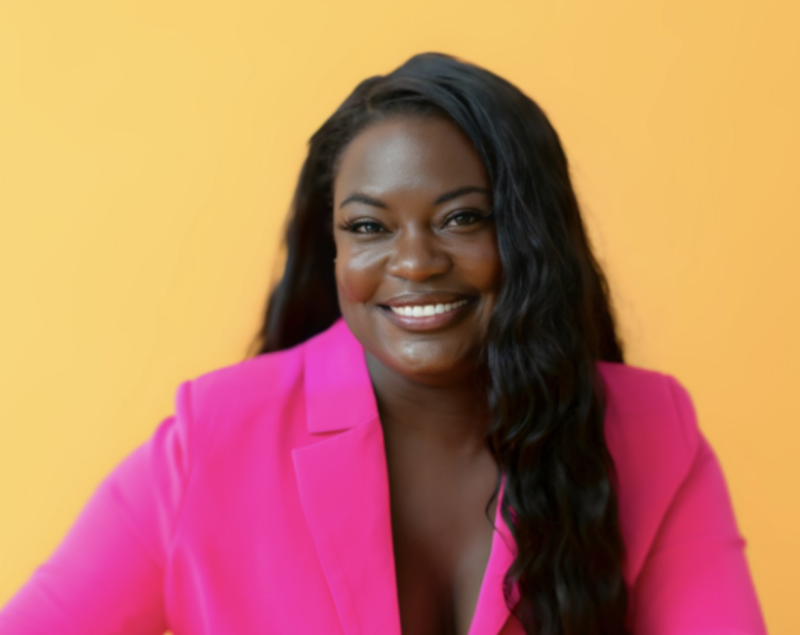 Raven Baxter poses for photo in a bright pink suit with a sunset orange background.