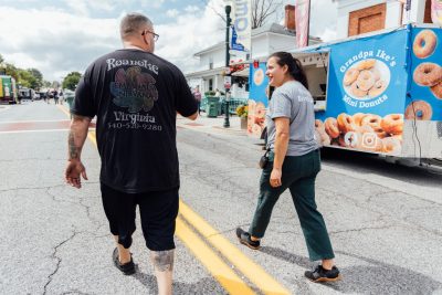 Elli walks with a food truck vendor up the middle of the street, food trucks line the street on either side. The two are talking and looking at each other as they walk.
