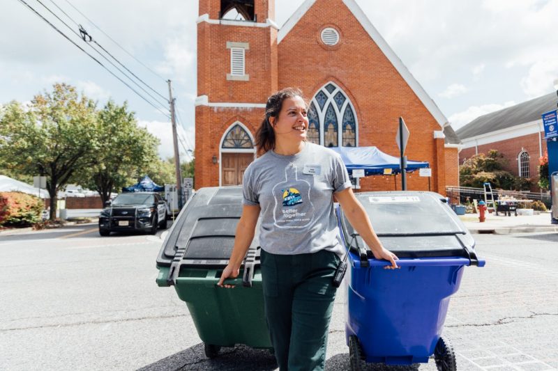 A woman pulls a large green recycling bin and a blue trash bin on wheels behind her as she walks across a street.